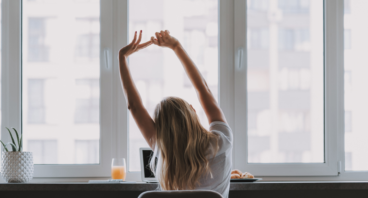 Someone stretching while working in front of a row of windows with a laptop, juice, and pastries