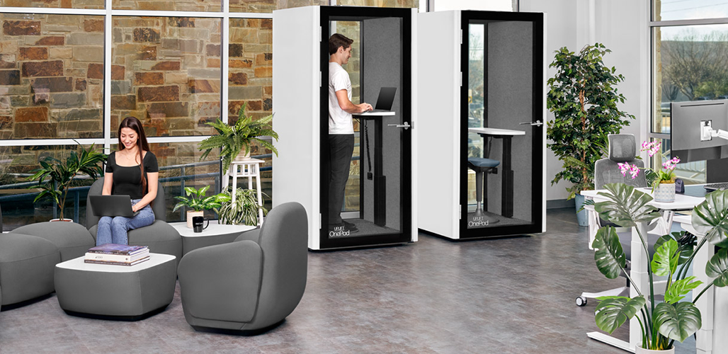 Open office with UPLIFT Desk's OnePod Office Phone Booth, where a woman takes a video call while a man works at a Standing Desk