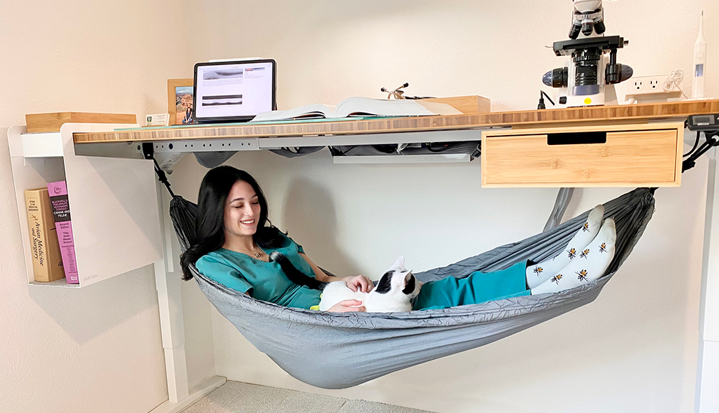 Woman laying in an under desk hammock by UPLIFT attached to a bamboo UPLIFT Desk featuring desk storage and accessories.