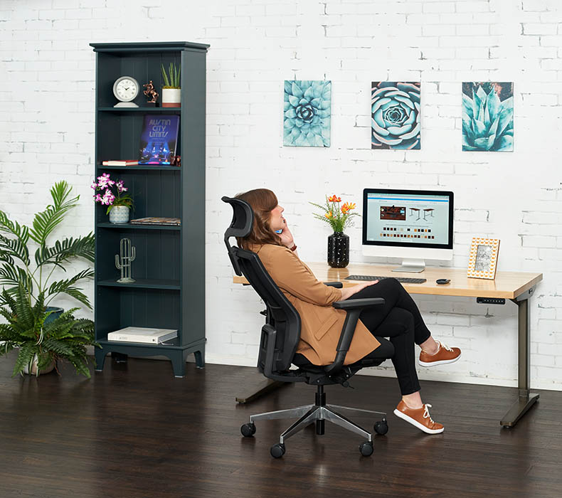 A person reclining in a black Envoke Ergonomic Chair beside her UPLIFT Standing Desk